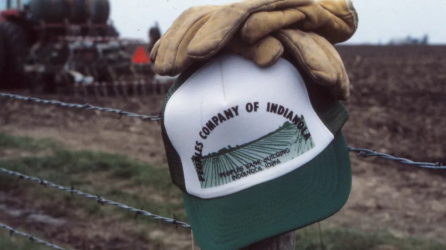 A vintage Peoples Company of Indianola cap and a pair of work gloves resting on a barbed-wire fence post, a planter working the field behind